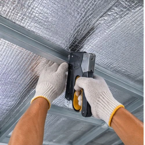 Worker wearing gloves uses a rivet gun to fasten a metal ceiling frame under reflective insulation panels.