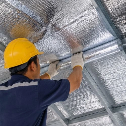 Pemasangan+Aluminium+foil+Single - Berkah Sentosa - Berkah Sentosa HVAC technician in a yellow hard hat and gloves installs reflective insulated duct in a metal ceiling framework.